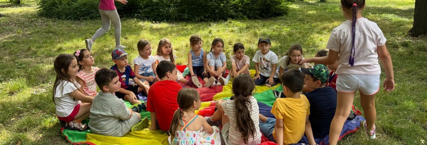 Copii asezati pe iarba in parc, la o activitate de grup, in tabara urbana de vara 5-7 ani.